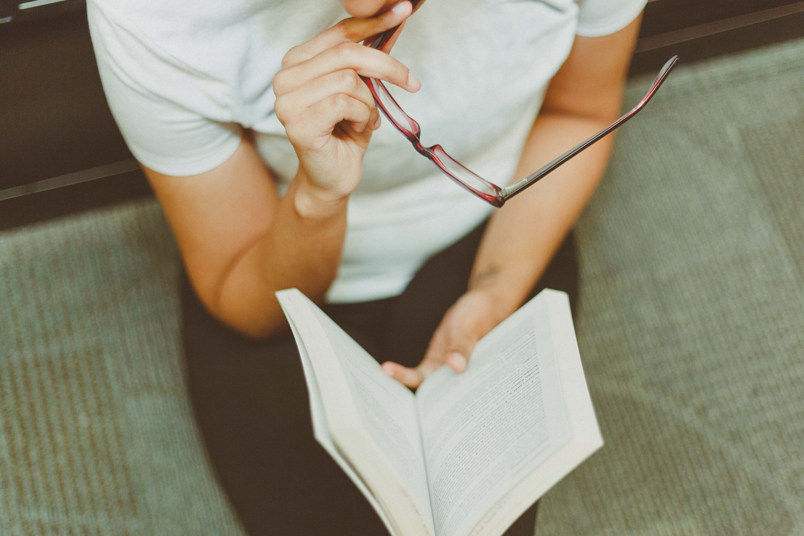 A photo of a woman holding reading glasses in her hand and reading a book.
